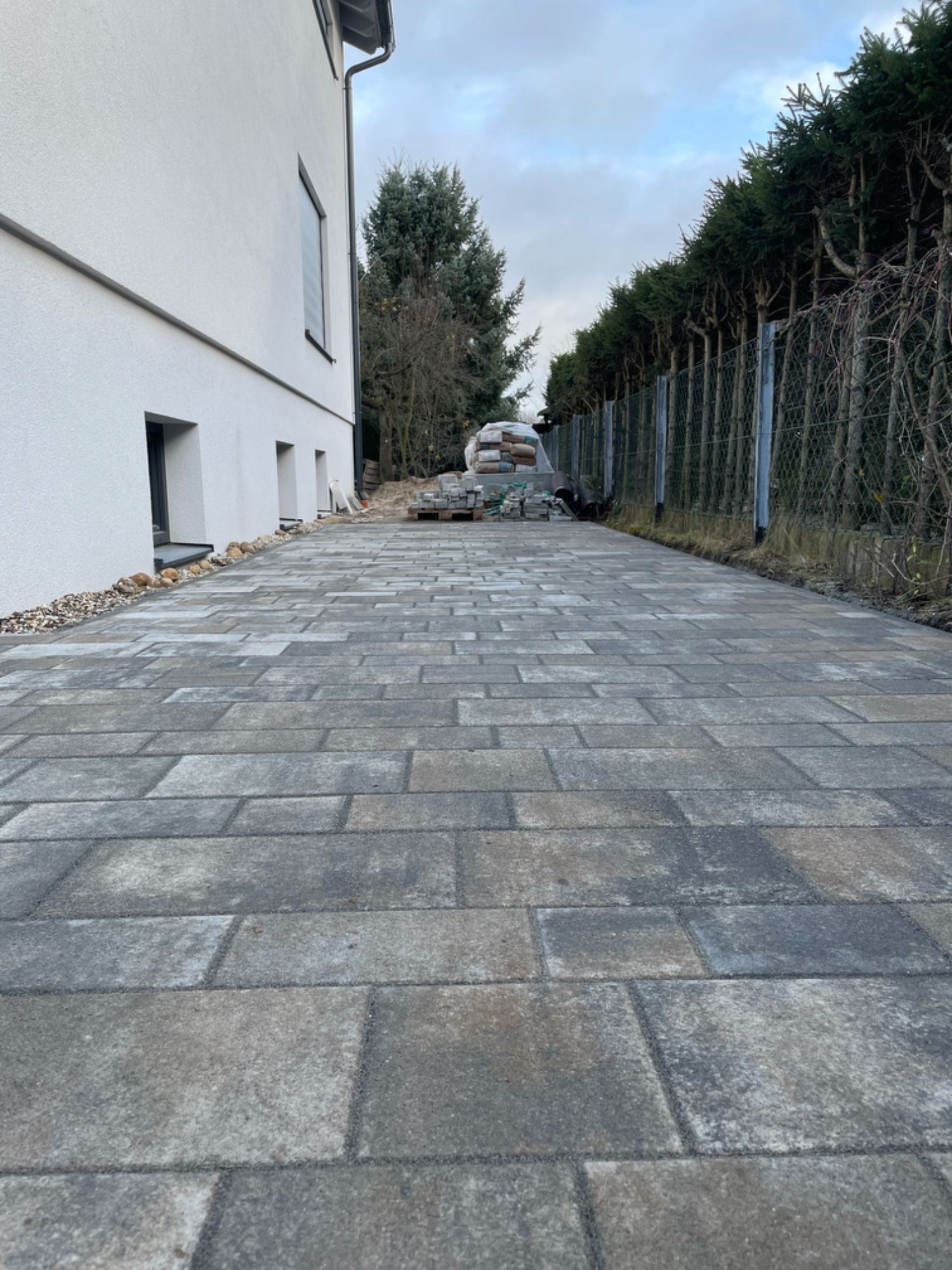 Gepflasterter Weg neben einem Gebäude, umgeben von Bäumen und einem Zaun.