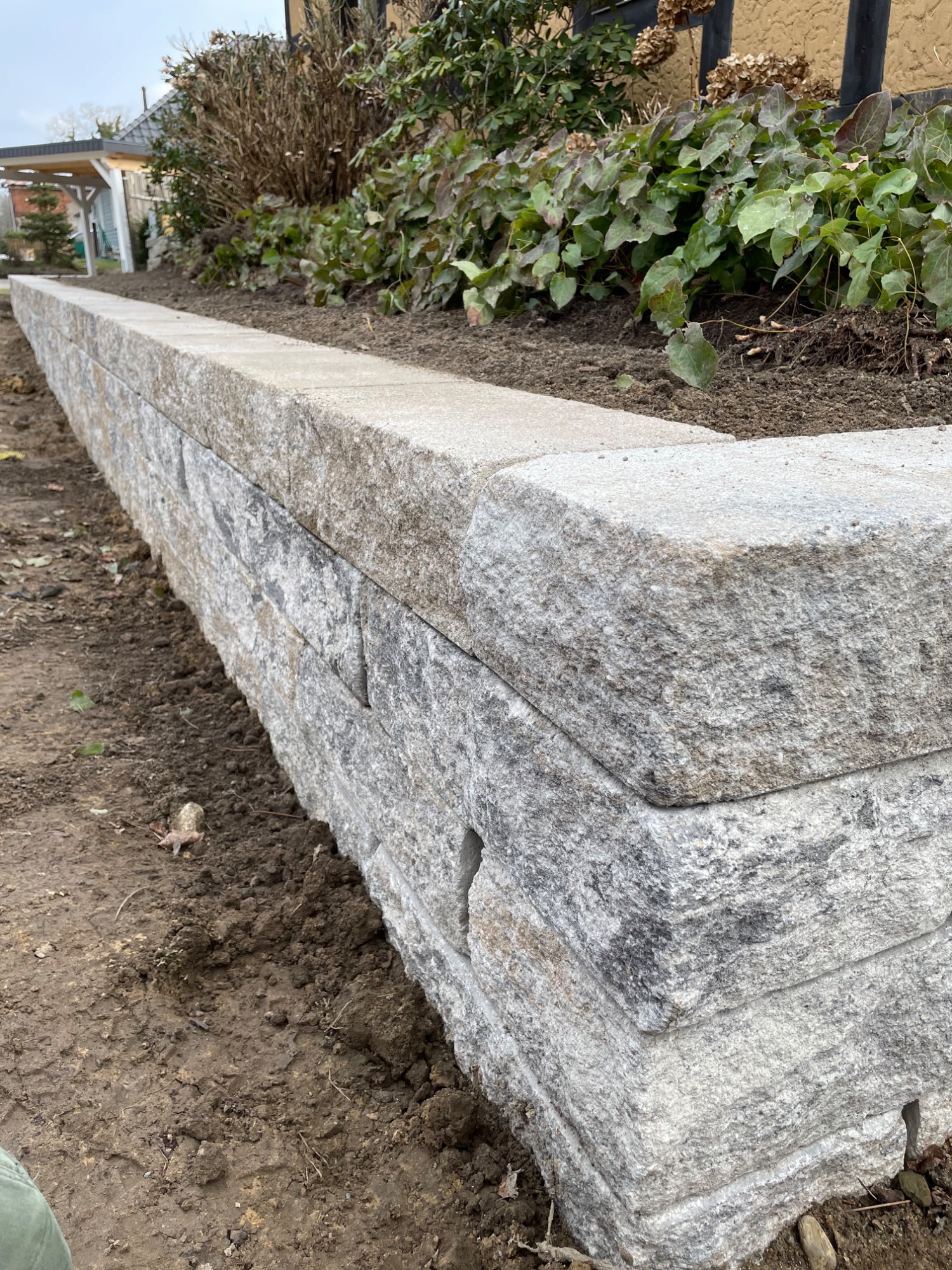 Ein niedriger Steinmauer aus grauem Naturstein, umgeben von Erde und Pflanzen.