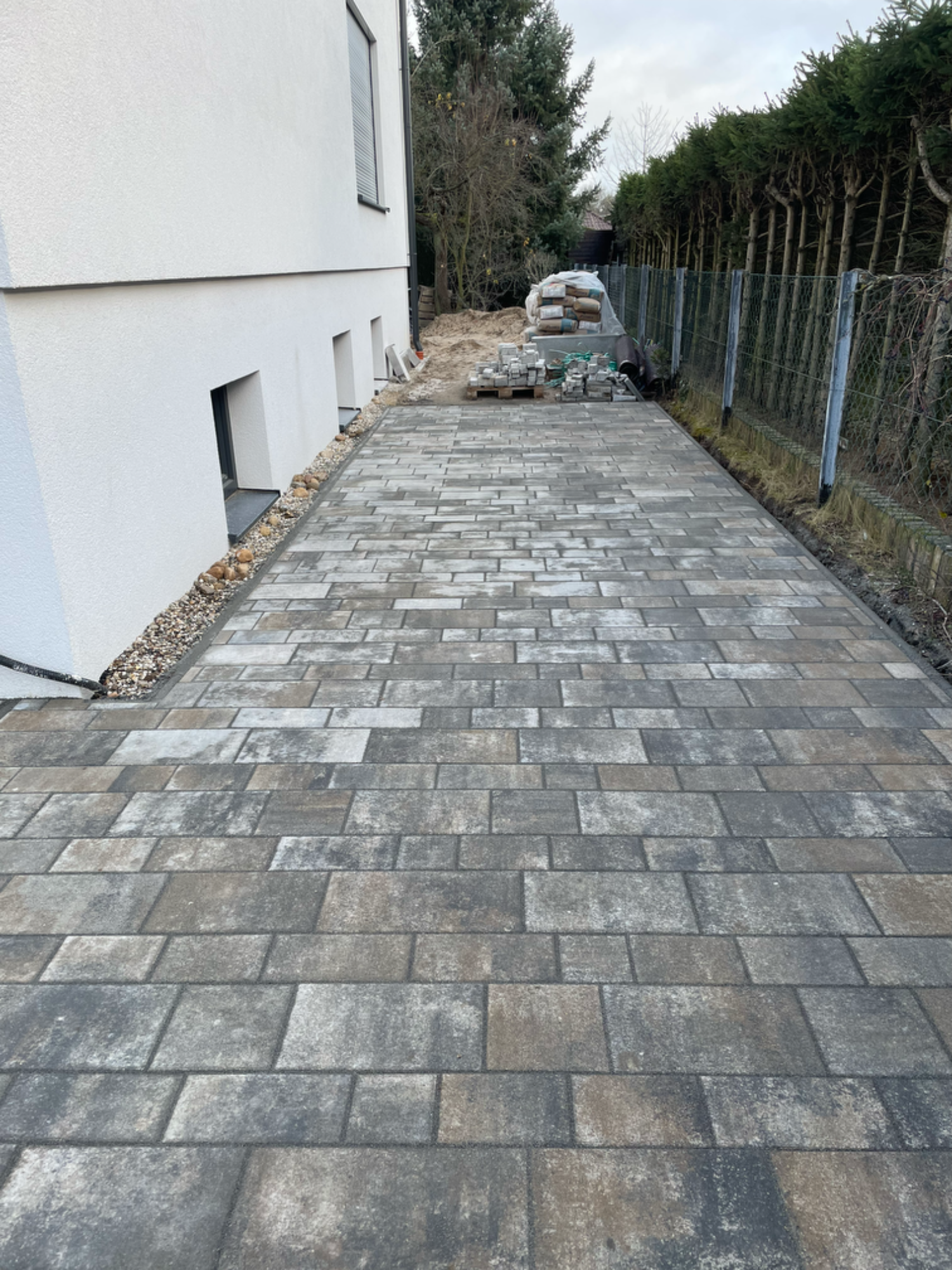 Gepflasterter Weg neben einem Gebäude, umgeben von Büschen und einem Zaun.