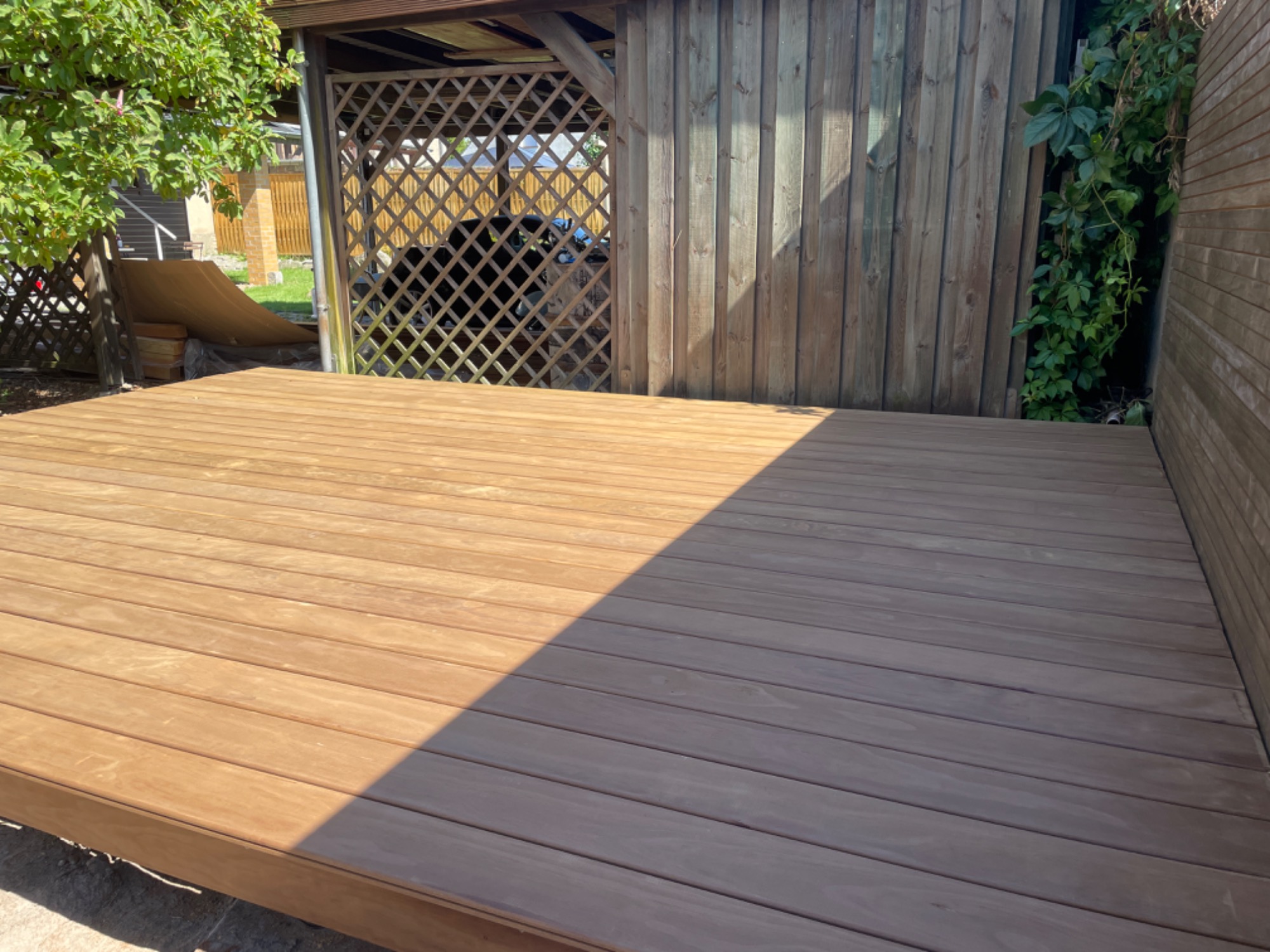 Holzterrasse vor einer Wand, teils im Schatten, umgeben von Pflanzen.