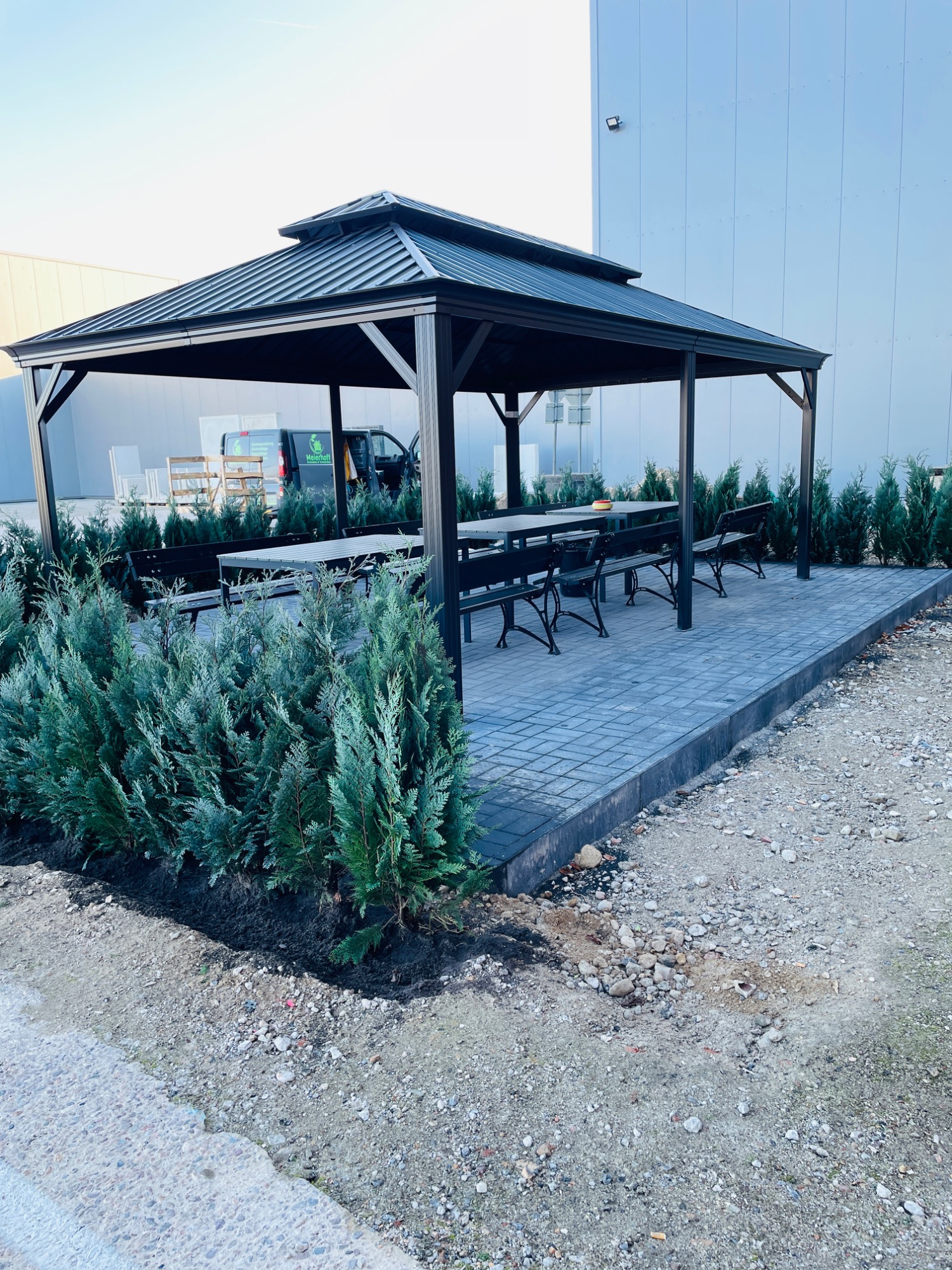 Pavillon mit Holztisch und Bänken, umgeben von grünen Sträuchern und gepflastertem Boden.