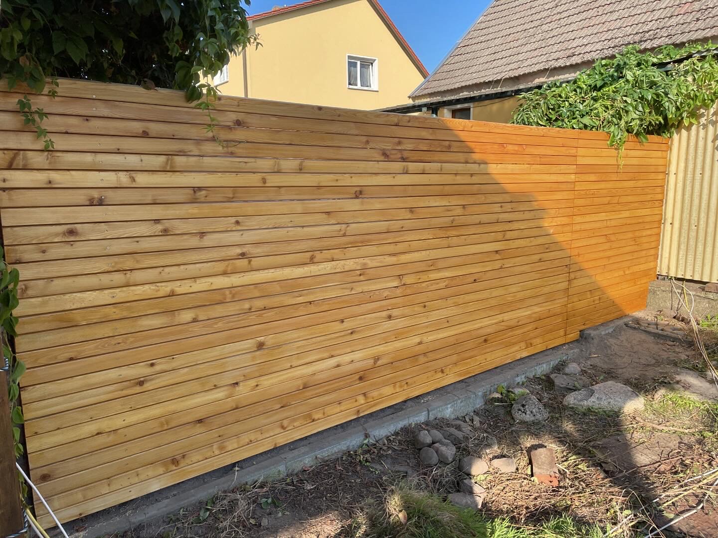 Holzsichtschutzzaun in verschiedenen Farbtönen, schräg angeordnet, umgeben von Pflanzen.