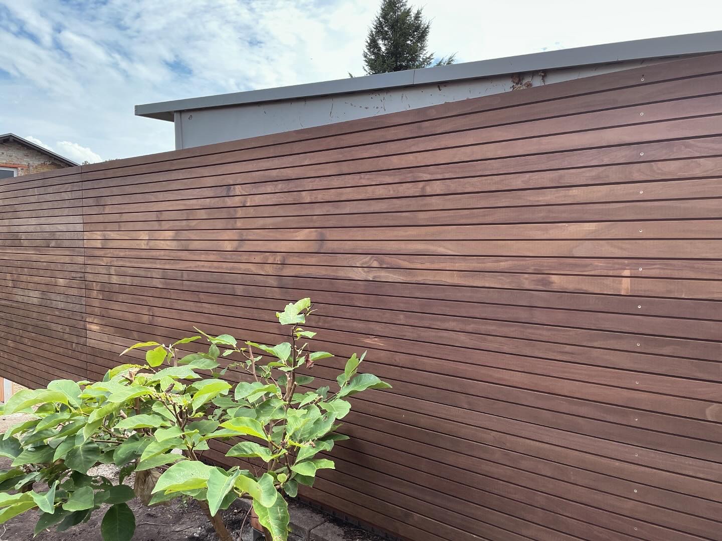 Holzlattenzaun mit horizontalen Elementen, im Hintergrund ein Gebäude und ein blühender Strauch.