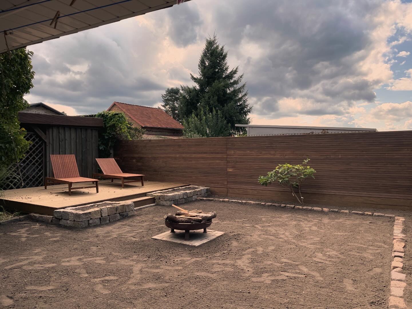 Gartenlandschaft mit zwei Liegestühlen, Feuerstelle und umgebenden Holzhecken.
